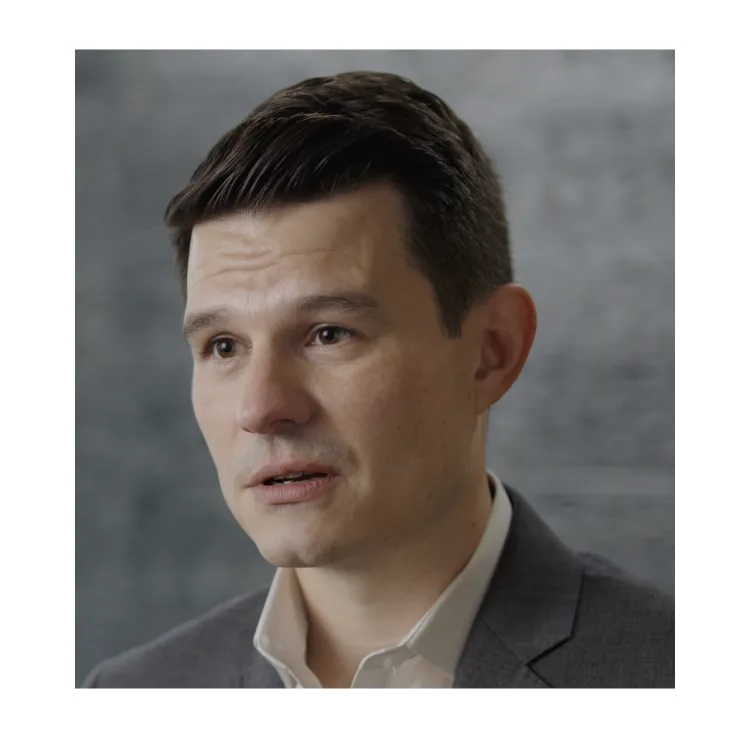 Portrait of a man with dark hair wearing a gray suit jacket and white shirt against a blurred gray background.